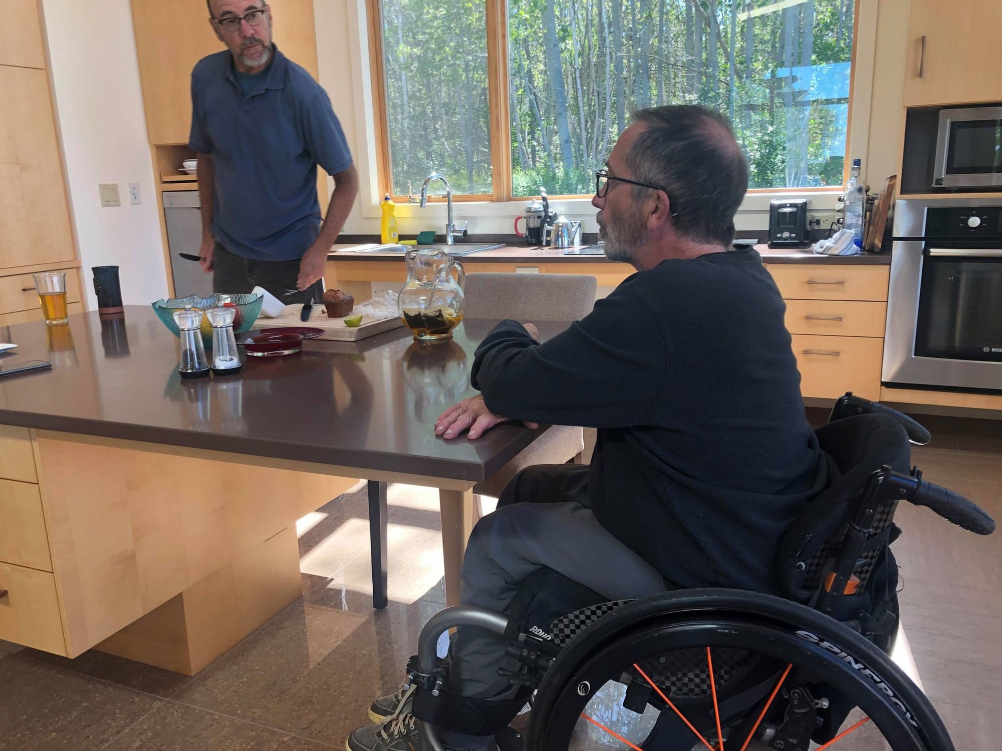 A man in a wheelchair sits at a kitchen island in an accessible home kitchen, talking with another man