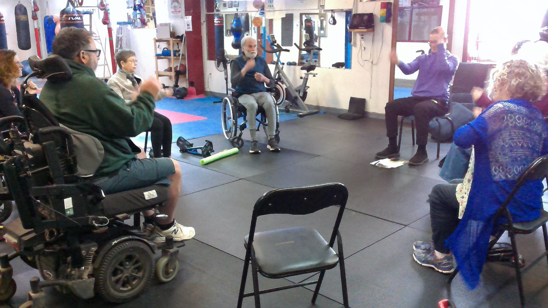 Participants in wheelchairs and chairs sit in a circle in a gym during a seated fitness session