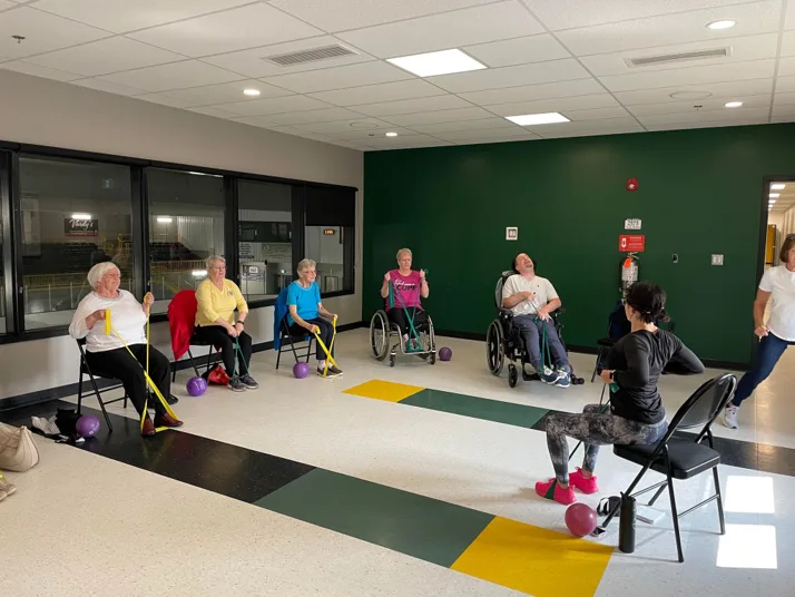 A seated fitness class: participants in chairs and wheelchairs exercise with resistance bands and balls while an instructor leads
